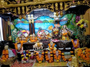 The image shows a vibrant altar with colorful decoration featuring multiple ornate deities adorned with floral garlands in the center. Surrounding the deities are various flowers, paintings, and sculptures, creating a festive and spiritual atmosphere. Bright marigold garlands embellish the surroundings, and there are depictions of religious figures or saints at the base. The backdrop includes a vividly painted mural with nature elements like trees and a peacock.