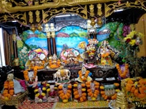 The image shows a vibrant altar with colorful decoration featuring multiple ornate deities adorned with floral garlands in the center. Surrounding the deities are various flowers, paintings, and sculptures, creating a festive and spiritual atmosphere. Bright marigold garlands embellish the surroundings, and there are depictions of religious figures or saints at the base. The backdrop includes a vividly painted mural with nature elements like trees and a peacock.