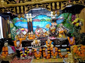 The image shows a vibrant altar with colorful decoration featuring multiple ornate deities adorned with floral garlands in the center. Surrounding the deities are various flowers, paintings, and sculptures, creating a festive and spiritual atmosphere. Bright marigold garlands embellish the surroundings, and there are depictions of religious figures or saints at the base. The backdrop includes a vividly painted mural with nature elements like trees and a peacock.