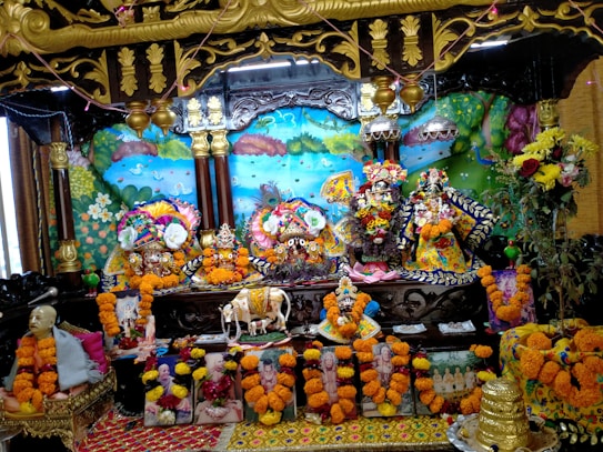 The image shows a vibrant altar with colorful decoration featuring multiple ornate deities adorned with floral garlands in the center. Surrounding the deities are various flowers, paintings, and sculptures, creating a festive and spiritual atmosphere. Bright marigold garlands embellish the surroundings, and there are depictions of religious figures or saints at the base. The backdrop includes a vividly painted mural with nature elements like trees and a peacock.