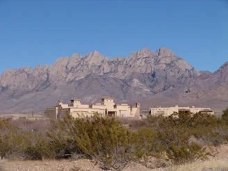 An adobe-style building is nestled at the base of a rugged mountain range, with sparse desert vegetation in the foreground and a clear blue sky above.