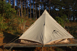 A beautifully lit premium canvas bell tent standing tall in a forest clearing at sunset, with warm interior lighting glowing through the canvas.