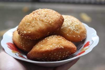 A plate with several golden-brown pastries topped with sesame seeds is being held. The pastries have a slightly crisp texture and are arranged on a white plate with a floral design.