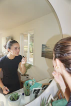 A confident woman applying makeup in front of a mirror with natural light.