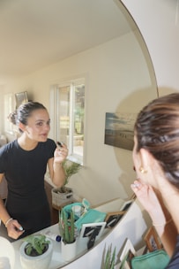 A person is applying makeup while looking into a mirror in a well-lit room. The reflection shows them holding a cosmetic item near their lips. On the table in front, there are potted plants, cosmetic products, and framed pictures. A window with blinds is visible in the background, letting in natural light.