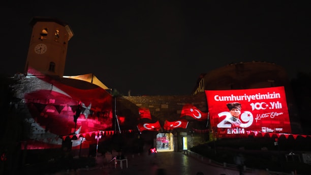 A nighttime scene featuring a historic building illuminated with red lights and projected images, including a large digital display celebrating the 100th anniversary of the republic on the right side. Turkish flags and decorations are visible, enhancing the festive atmosphere, with a clock tower in the background.