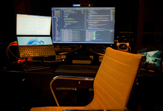 A computer setup featuring multiple screens with a focus on software development. The central monitor displays code in a dark-themed editor. A laptop and an additional monitor are arranged on a desk. The environment is dimly lit, and a swivel chair sits empty in front of the desk.
