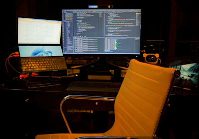 A computer setup featuring multiple screens with a focus on software development. The central monitor displays code in a dark-themed editor. A laptop and an additional monitor are arranged on a desk. The environment is dimly lit, and a swivel chair sits empty in front of the desk.