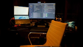 A computer setup featuring multiple screens with a focus on software development. The central monitor displays code in a dark-themed editor. A laptop and an additional monitor are arranged on a desk. The environment is dimly lit, and a swivel chair sits empty in front of the desk.