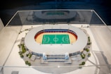 A detailed architectural model of a sports stadium is encased in a clear protective case. The stadium features a central green field marked for sports games, surrounded by a red and blue seating area. The exterior of the structure is adorned with decorative columns and small trees are planted around the model, simulating a realistic environment.