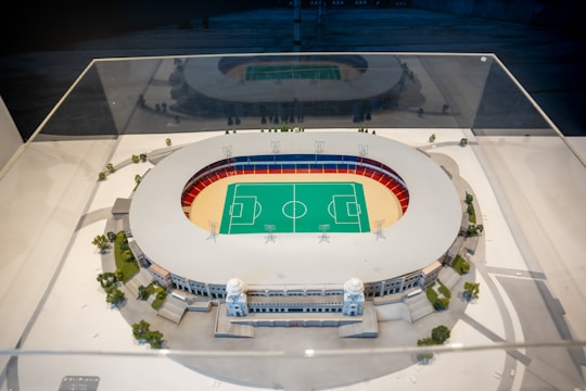 A detailed architectural model of a sports stadium is encased in a clear protective case. The stadium features a central green field marked for sports games, surrounded by a red and blue seating area. The exterior of the structure is adorned with decorative columns and small trees are planted around the model, simulating a realistic environment.
