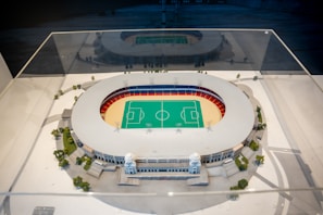 A detailed architectural model of a sports stadium is encased in a clear protective case. The stadium features a central green field marked for sports games, surrounded by a red and blue seating area. The exterior of the structure is adorned with decorative columns and small trees are planted around the model, simulating a realistic environment.