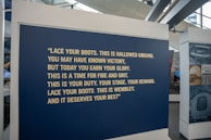 A large blue panel featuring a motivational quote in bold, yellow text is displayed at an exhibition. The quote encourages resilience and pride, specifically referencing Wembley. The area surrounding the panel includes other display stands and large windows.