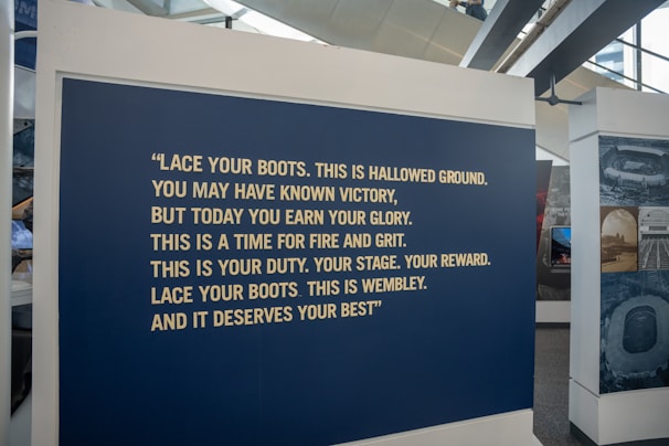 A large blue panel featuring a motivational quote in bold, yellow text is displayed at an exhibition. The quote encourages resilience and pride, specifically referencing Wembley. The area surrounding the panel includes other display stands and large windows.