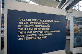 A large blue panel featuring a motivational quote in bold, yellow text is displayed at an exhibition. The quote encourages resilience and pride, specifically referencing Wembley. The area surrounding the panel includes other display stands and large windows.