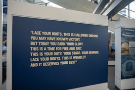 A large blue panel featuring a motivational quote in bold, yellow text is displayed at an exhibition. The quote encourages resilience and pride, specifically referencing Wembley. The area surrounding the panel includes other display stands and large windows.