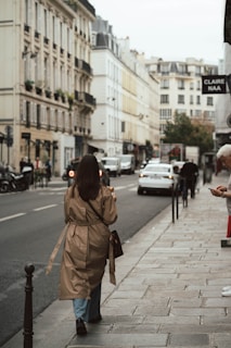 A sunlit street scene featuring a woman in a chic trench coat and classic ballet flats walking past a café.
