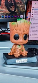 A cute figurine of a character with a large head and wide eyes sitting on a desk. The figurine is from the 'Guardians of the Galaxy' series and is positioned in front of a laptop. Behind the laptop, there is a computer monitor displaying various application icons.