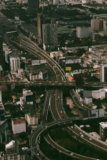 An aerial view of an urban area featuring a complex network of highways and overpasses surrounded by tall buildings. The roads are bustling with vehicles, and residential and commercial structures are densely packed around the highways.