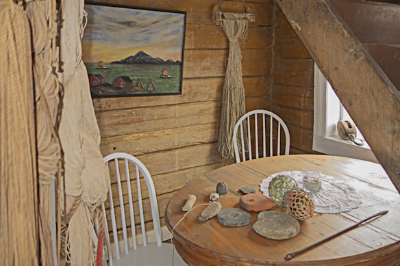 A rustic wooden room with a round table decorated with miscellaneous items like stones and a decorative orb. Two white chairs with spindles are placed against the log walls. A painting of a rural landscape featuring mountains and small houses is hung on the wall. Thick ropes are hanging beside the chair, and there is a small window on the right side.