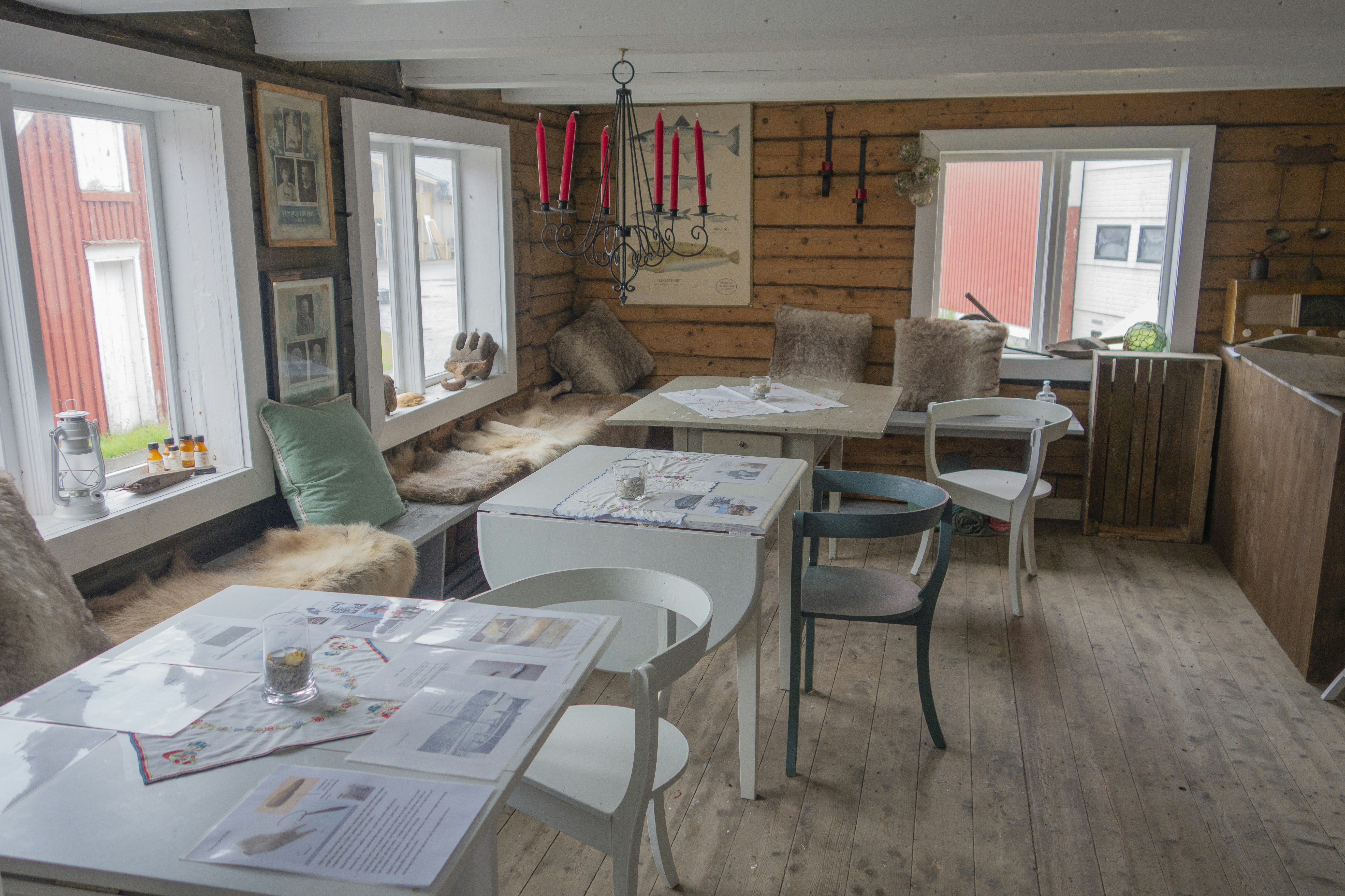 A cozy, rustic room with wooden walls and floor, featuring several white and light-colored chairs around tables covered with papers and menus. Soft fur throws and cushions line a bench by the windows, under a black chandelier with red candles. Various decorative items, including framed pictures and small objects, are visible around the space, creating a warm and inviting atmosphere.
