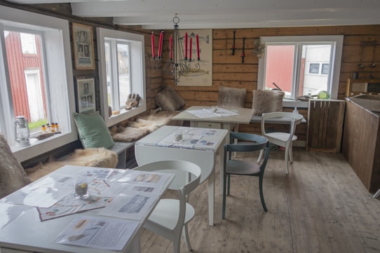 A cozy, rustic room with wooden walls and floor, featuring several white and light-colored chairs around tables covered with papers and menus. Soft fur throws and cushions line a bench by the windows, under a black chandelier with red candles. Various decorative items, including framed pictures and small objects, are visible around the space, creating a warm and inviting atmosphere.