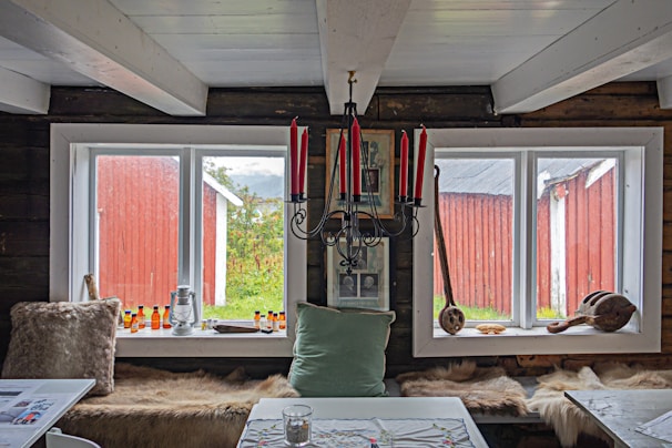 A rustic interior features two large windows framed in white, offering a view of a red wooden building and greenery outside. The room is decorated with a black chandelier holding red candles, various framed pictures, and a long seat with animal fur throw pillows. A light green pillow and a tabletop with a fabric cover also appear, along with bottles and wooden objects as decoration.