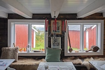 A rustic interior features two large windows framed in white, offering a view of a red wooden building and greenery outside. The room is decorated with a black chandelier holding red candles, various framed pictures, and a long seat with animal fur throw pillows. A light green pillow and a tabletop with a fabric cover also appear, along with bottles and wooden objects as decoration.