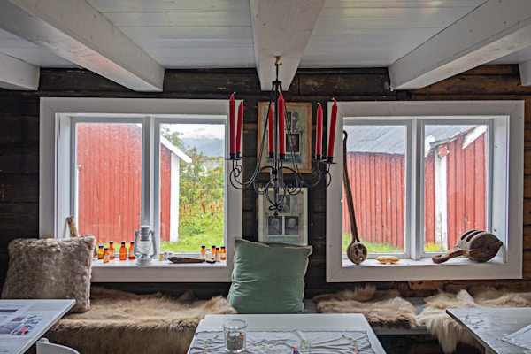 A rustic interior features two large windows framed in white, offering a view of a red wooden building and greenery outside. The room is decorated with a black chandelier holding red candles, various framed pictures, and a long seat with animal fur throw pillows. A light green pillow and a tabletop with a fabric cover also appear, along with bottles and wooden objects as decoration.