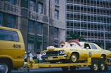 A yellow vehicle resembling a police car is on a flatbed trailer, with a gigantic pair of eyes fixed on its hood, giving it an animated appearance. Adjacent to this, a yellow van numbered 409 is part of the towing setup. The backdrop features a multi-story building with numerous windows, and a group of people stands on the sidewalk, observing the scene. The atmosphere is vibrant and quirky.