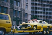 A yellow vehicle resembling a police car is on a flatbed trailer, with a gigantic pair of eyes fixed on its hood, giving it an animated appearance. Adjacent to this, a yellow van numbered 409 is part of the towing setup. The backdrop features a multi-story building with numerous windows, and a group of people stands on the sidewalk, observing the scene. The atmosphere is vibrant and quirky.