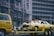 A yellow vehicle resembling a police car is on a flatbed trailer, with a gigantic pair of eyes fixed on its hood, giving it an animated appearance. Adjacent to this, a yellow van numbered 409 is part of the towing setup. The backdrop features a multi-story building with numerous windows, and a group of people stands on the sidewalk, observing the scene. The atmosphere is vibrant and quirky.