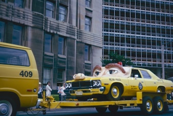 A yellow vehicle resembling a police car is on a flatbed trailer, with a gigantic pair of eyes fixed on its hood, giving it an animated appearance. Adjacent to this, a yellow van numbered 409 is part of the towing setup. The backdrop features a multi-story building with numerous windows, and a group of people stands on the sidewalk, observing the scene. The atmosphere is vibrant and quirky.