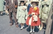 Two young girls stand side by side, dressed in formal outfits with hats. The girl on the left wears a pastel green suit with pink flowers pinned to her lapel, while the girl on the right is in a bright red coat adorned with a large yellow flower. Both girls hold small handbags and are surrounded by adults whose clothing is also visible.