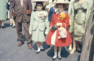 Two young girls stand side by side, dressed in formal outfits with hats. The girl on the left wears a pastel green suit with pink flowers pinned to her lapel, while the girl on the right is in a bright red coat adorned with a large yellow flower. Both girls hold small handbags and are surrounded by adults whose clothing is also visible.