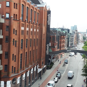 A street scene featuring historic red-brick buildings with the word 'stilwerk' visible on banners. The buildings have large arched windows and there is a modern glass bridge connecting two structures. The street is cobbled with parked cars, bikes, and a few pedestrians. The area looks urban and is likely part of a cityscape.