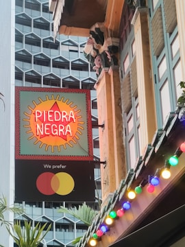 A vibrant street scene featuring a neon sign with 'Piedra Negra' in bold letters. The sign is mounted on a building with colorful decorative elements and surrounded by strings of colorful lights. In the background, there is a modern building with a geometric pattern.