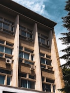 A multi-story building facade with rows of windows and beige concrete panels. Two air conditioning units are visible attached to the exterior. The sky is partly cloudy, and a tree is seen on the right.