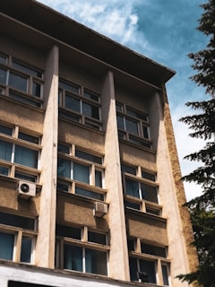 A multi-story building facade with rows of windows and beige concrete panels. Two air conditioning units are visible attached to the exterior. The sky is partly cloudy, and a tree is seen on the right.