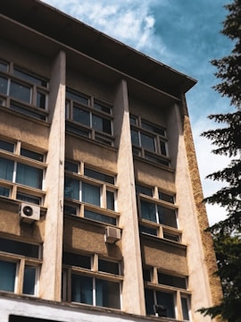 A multi-story building facade with rows of windows and beige concrete panels. Two air conditioning units are visible attached to the exterior. The sky is partly cloudy, and a tree is seen on the right.