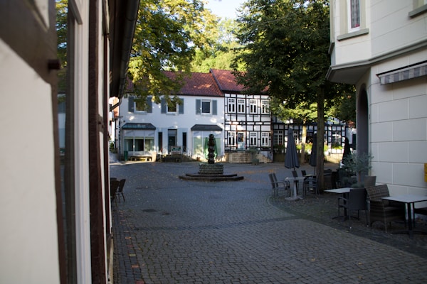 The image depicts a quaint, cobblestone street in a small town with traditional, half-timbered buildings. The area appears to be a small courtyard or plaza with empty outdoor seating areas. Sunlight filters through the leaves of the trees, casting dappled shadows on the pavement.