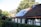 A charming house with a sloped roof featuring solar panels and some moss-covered tiles. Surrounding the property is a lush green hedge, and mature trees provide a natural backdrop.