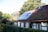 A charming house with a sloped roof featuring solar panels and some moss-covered tiles. Surrounding the property is a lush green hedge, and mature trees provide a natural backdrop.