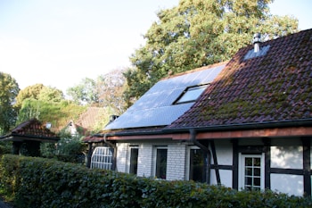 A charming house with a sloped roof featuring solar panels and some moss-covered tiles. Surrounding the property is a lush green hedge, and mature trees provide a natural backdrop.