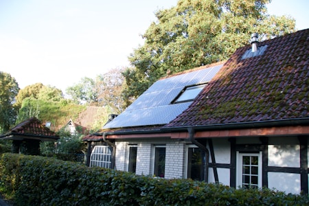 A charming house with a sloped roof featuring solar panels and some moss-covered tiles. Surrounding the property is a lush green hedge, and mature trees provide a natural backdrop.