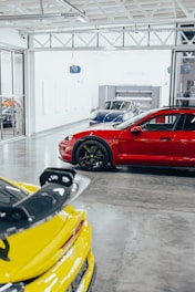 A spacious, modern garage featuring three luxury sports cars in the foreground. The vehicles include a vibrant red car with sleek black wheels, a yellow car with a large spoiler, and a shiny blue car in the background. The floor is polished concrete and the garage is well-lit with natural and artificial lights, giving it a clean and stylish appearance.