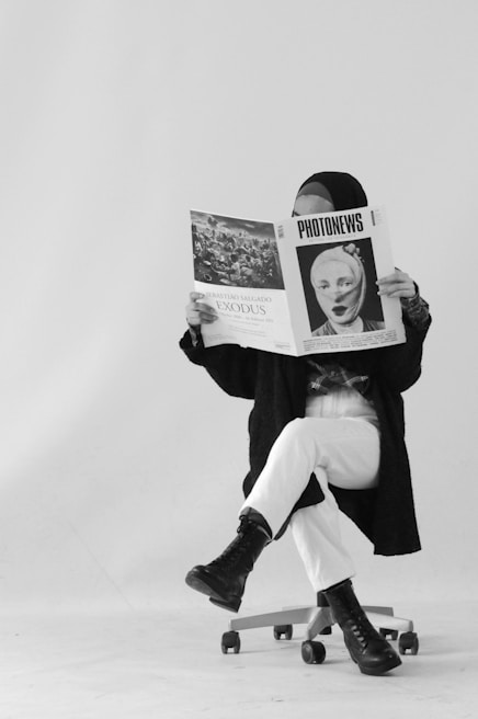 A person sits on a swivel chair, legs crossed, reading a large newspaper titled 'PHOTONEWS' with a black-and-white image on the cover. They are wearing a hijab, a dark coat, white pants, and black boots. The background is plain and white, adding an artistic, minimalistic feel.
