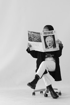 A person sits on a swivel chair, legs crossed, reading a large newspaper titled 'PHOTONEWS' with a black-and-white image on the cover. They are wearing a hijab, a dark coat, white pants, and black boots. The background is plain and white, adding an artistic, minimalistic feel.