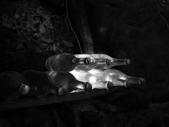 Several wine bottles are laying horizontally on a wooden rack against a rocky, dimly lit background. Light subtly illuminates the transparent, empty bottles, casting gentle reflections.