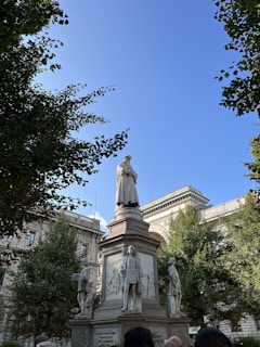 A statue stands prominently in a park area surrounded by trees and historic buildings. The statue is on a pedestal and features figures, with 'Leonardo' inscribed on it. People can be seen at the base of the statue, suggesting a public place.